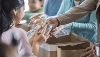 Young woman receives a canned food item from food bank. An unrecognizable volunteer hands her the canned food item.